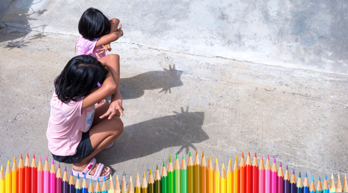 Due bambine accovacciate giocano con le ombre simulando dei gatti. nella parte bassa dell'immagine si vedono delle matite colorate in gradazione.