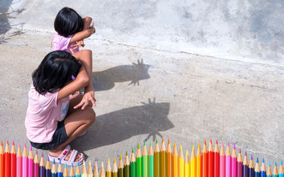 Due bambine accovacciate giocano con le ombre simulando dei gatti. nella parte bassa dell'immagine si vedono delle matite colorate in gradazione.